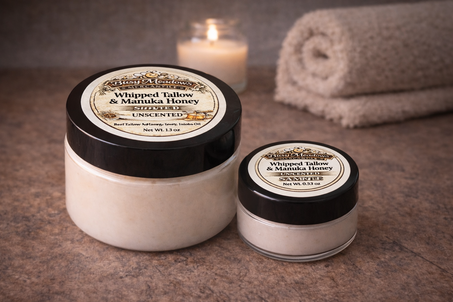Two jars of whipped tallow and manuka honey on a textured surface with a candle and towel in the background.